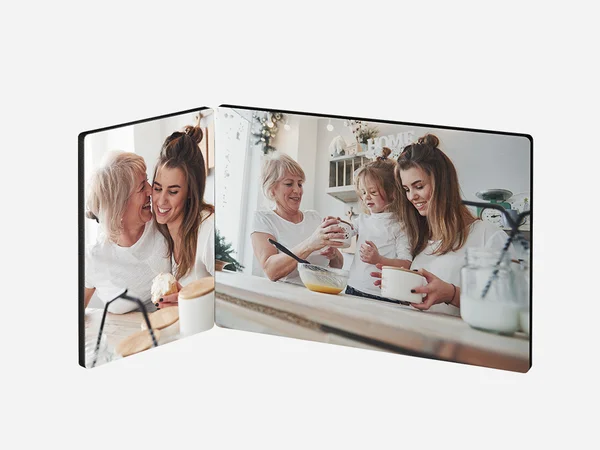 Ein zweiteiliger Fotoaufsteller zeigt zwei fröhliche Szenen in einer Küche. Auf der linken Seite umarmen sich eine ältere Frau mit weißen Haaren und eine junge Frau mit braunen Haaren und lachen. Sie stehen an einem Tisch mit einer Glasflasche.