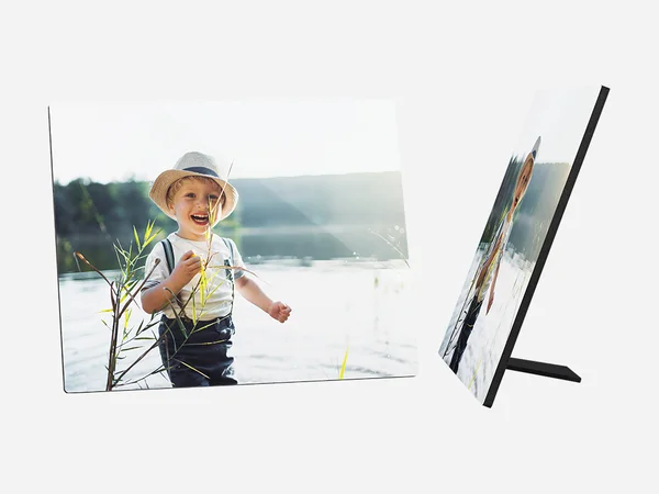 Ein quadratischer Fotoaufsteller zeigt ein fröhliches kleines Kind, das am Ufer eines Sees oder Flusses steht. Das Kind hat blonde Haare und trägt einen hellen Hut, ein weißes T-Shirt mit einem bunten Aufdruck und dunkle Latzhosen. 